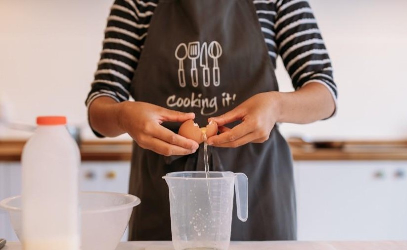 Person wearing a 'Cooking It' apron cracking an egg into a jug 